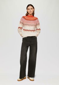 Strickpullover mit rot-rosa Zickzackmuster auf crèmefarbenem Grund, kombiniert mit schwarzer weitgeschnittener Jeans und braunen Schuhen mit weißen Akzenten.