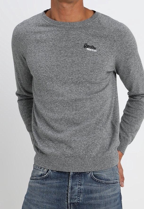 Homme portant un pull gris ajusté en maille avec un petit logo noir sur le côté gauche de la poitrine, associé à un jean en denim bleu.