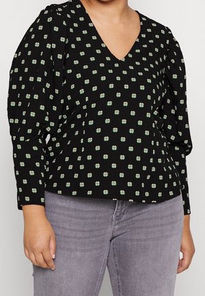 Blouse noire à col en V avec manches longues bouffantes et petit motif floral blanc, portée avec un jean gris clair.