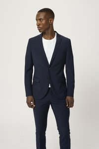 Costume bleu marine avec blazer à bouton unique et pantalon assorti, associé à un t-shirt blanc. Tissu lisse avec une coupe ajustée, poches discrètes.