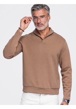Mand med gråt hår og skæg justerer klare briller, iført en brun pullover og hvide bukser, mod en ensfarvet lys baggrund.