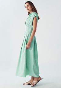 Mätovo zelená maxi šaty s krátkymi, štruktúrovanými rukávmi, s vertikálnymi skladmi a vysokým výstrihom. V kombinácii s bielymi sandálmi na remienky.