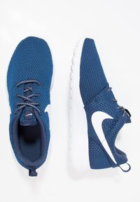 Siniset verkkokangasta urheilukengät, joissa on valkoiset kumipohjat ja yksityiskohtia. Ominaisuuksiin kuuluu vahvat nauhat ja kantapäässä oleva lenkki helppoa pukemista varten.