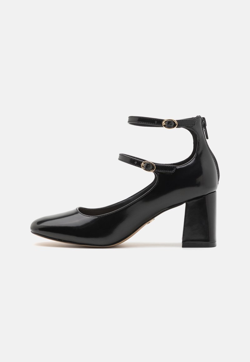 Chaussure Mary Jane en cuir verni noir avec deux fines brides, boucles dorées, talon bloc et fermeture éclair arrière sur fond gris clair.