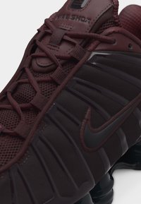 Gros plan sur une chaussure Nike Shox grenat et noire avec un laçage détaillé, une tige en mesh et le logo Nike visible sur le côté.