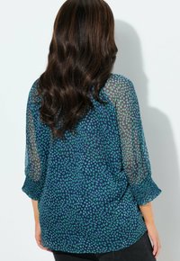 Modrá a zelená blůza se vzorem, průsvitnými rukávy, kulatým výstřihem a natrhanými manžetami. Obsahuje malé srdíčkové tvary v posunutém uspořádání.