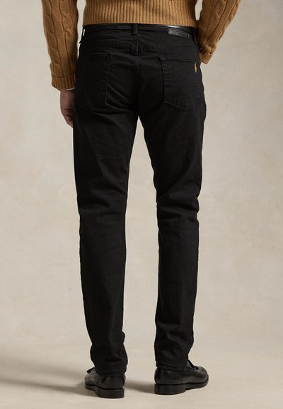 Férfi fekete farmert, barna fonott mintás pulóvert és fekete bőr cipőt visel, háttal áll a kamerának egy semleges háttér előtt.