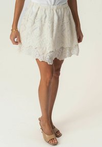 Jupe patineuse en dentelle blanche avec des motifs floraux, ourlet festonné et taille élastique. Portée avec des talons ouverts beige.