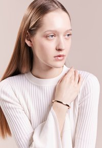 Auksinės spalvos metalinė apyrankė, dėvima ant modelio riešo, su lygiu, atspindinčiu paviršiumi ir paprastu, elegantišku dizainu. Modelis dėvi ribiuotą baltą marškinėlį.