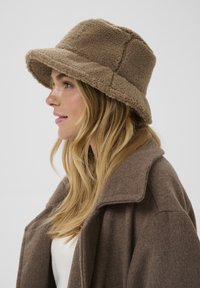 Brązowy kapelusz typu bucket z polaru z szerokim rondem, wykonany z teksturowanej tkaniny z pikowanymi detalami, noszony z beżowym płaszczem.
