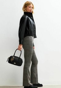 Femme portant une veste en cuir noire, un pantalon à carreaux et des chaussures noires, tenant un sac à main noir avec un pendentif en forme de cœur en imprimé léopard.