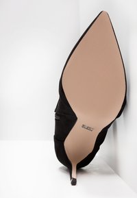 Chaussure à talon aiguille noire à bout pointu vue de la semelle, avec une semelle beige et une petite étiquette métallique de la marque.