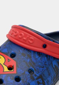 Blue and black Crocs with a red strap featuring a Superman logo. The surface displays comic-style graphics and ventilation holes.