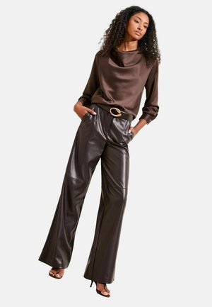 Kobieta o kręconych włosach, ubrana w brązową satynową bluzkę i ciemne skórzane spodnie z szerokimi nogawkami i wysokim stanem, ozdobione paskiem z złotą klamrą, na obcasach.