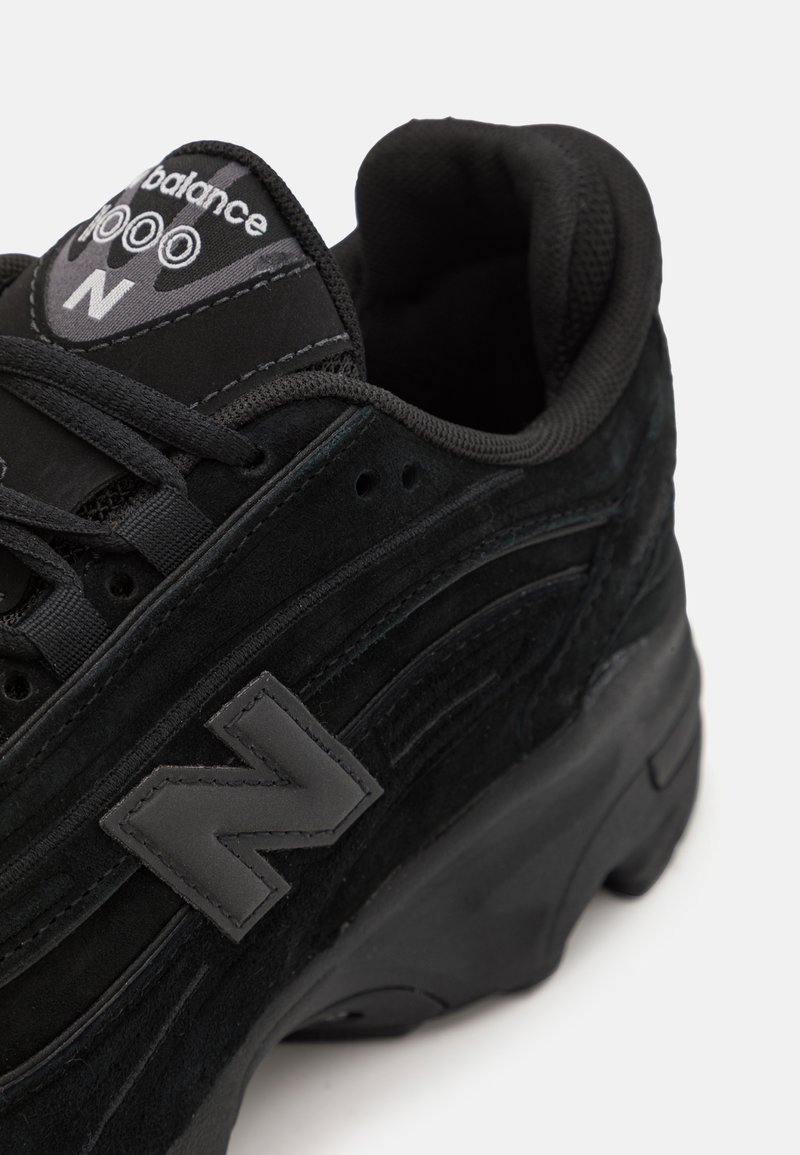 Zapatillas de ante negro con paneles laterales texturizados, que cuentan con un collar acolchado, diseño de cordones y un logo negro prominente en la lengüeta.