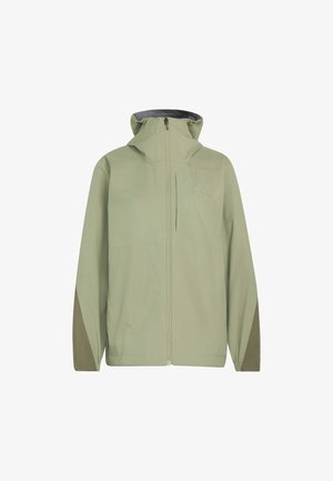 Mujer con chaqueta con capucha de color verde claro, pantalones negros y zapatillas beige caminando frente a un fondo claro y liso.
