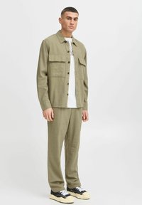 Veste chemise vert olive avec des boutons sur le devant et deux poches poitrine, assortie à un pantalon fuselé et des baskets noires avec des semelles blanches.