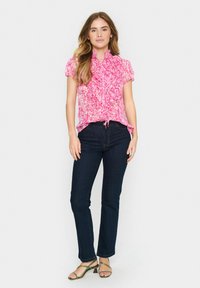 Pink, patterned blouse with ruffled design, short sleeves, and a tie at the waist, paired with dark denim jeans and green strappy sandals.