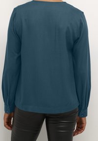 Blouse teal à manches longues avec un col arrondi, texture lisse, poignets boutonnés et une coupe légèrement ample. Portez-la avec un pantalon noir ajusté.