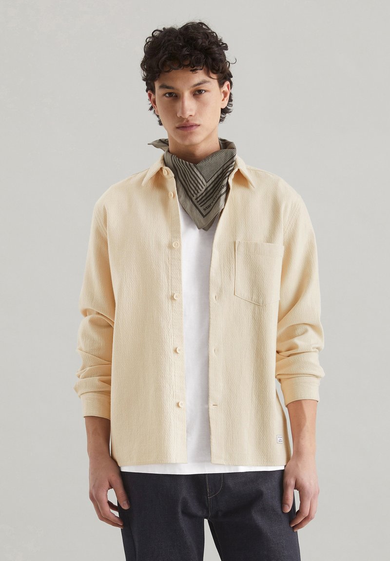 Cream-colored waffle-textured shirt with long sleeves, button-up front, and a single chest pocket, worn over a white t-shirt.