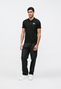 Svart poloshirt med en logo, kombinert med svarte, løstsittende bukser og hvite joggesko. Mann som står foran en nøytral bakgrunn.