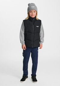 Schwarze Pufferweste über einem grauen Sweatshirt, marineblaue Trainingshose mit weißen Seitestreifen und einer grauen gerippten Mütze, kombiniert mit schwarzen Sneakers.