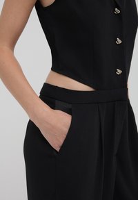 Haut sans manches noir avec des boutons et un design découpé, associé à un pantalon noir taille haute doté de poches latérales et d'une texture lisse.