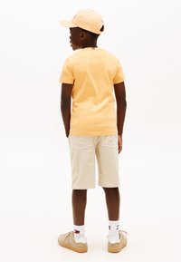 Αγόρι που κοιτάζει προς τα πίσω, φοράει ανοιχτό πορτοκαλί καπέλο και μπλουζάκι, μπεζ σορτς cargo, άσπρες κάλτσες με κόκκινη και μπλε ρίγα, και καφέ αθλητικά παπούτσια.