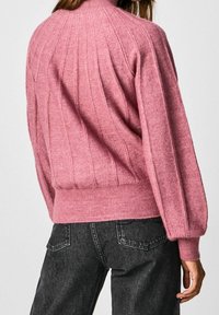 Růžový pletený svetr s vertikálním žebrováním, vysokým límcem a nabíranými rukávy; kombinovaný s tmavými džíny. Viditelně měkká textura.