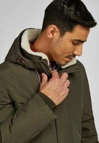 Olijfgroen jack met een gevoerde capuchon van fleece, ritssluiting en trekkoordaccenten. Heeft een gestructureerd buitenmateriaal en geribbelde manchetten.