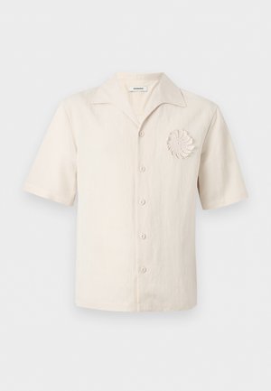 Mann mit lockigem Haar, der ein kurzärmeliges cremefarbenes Hemd mit Blumenmuster, schwarze weit geschnittene Hosen und schwarze Schuhe trägt, steht mit den Händen in den Taschen.