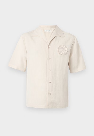 Πουκάμισο από λινό σε κρεμ απόχρωση με κοντά μανίκια, γιακά με εγκοπή, κουμπιά μπροστά και διακοσμητικό σπιράλ πλέγμα στο αριστερό μέρος του στήθους.