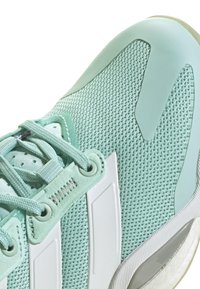 Zapatilla deportiva de color verde claro con parte superior de malla, detalles lisos en blanco, cordones texturizados y suela de goma con un talón ligeramente elevado.