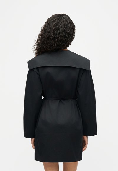 Γυναίκα με σγουρά μαλλιά, φοράει μαύρο παλτό με μακριά μανίκια, φαρδύ γιακά και ζώνη, με φορά προς τα πίσω σε απλό λευκό φόντο.