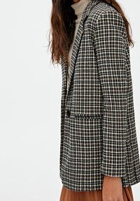 Blazer a scacchi in bianco e nero con un solo bottone, rever a V, tasche laterali e spalle strutturate in un tessuto testurizzato.