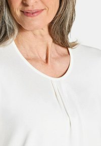 Weißes Oberteil mit Rundhalsausschnitt, geplisster Detailgestaltung und weicher, glatter Textur. Es hat eine subtile, hellfarbige Borte entlang des Ausschnitts.