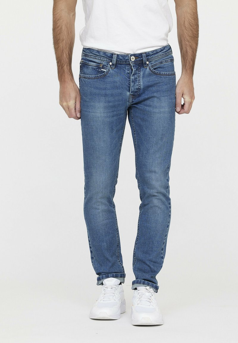 Jean en denim bleu clair, coupe slim, avec cinq poches, coutures contrastées et fermeture à bouton. Porté avec un t-shirt blanc uni.