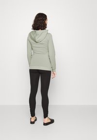 Un sweat à capuche zippé vert clair avec une capuche, une coupe ajustée et un ourlet côtelé, associé à des leggings noirs et des chaussures noires à enfiler.