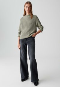Maglione verde a coste con colletto alto, abbinato a jeans grigi scuri a gamba larga. Modella in piedi contro uno sfondo neutro, con le mani in tasca.