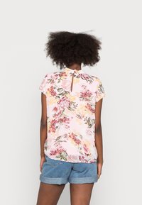 Kortärmad blus i ljusrosa med ett blommönster som har rosor och gula accenter; design med nyckelhål bak; slät tygtextur.