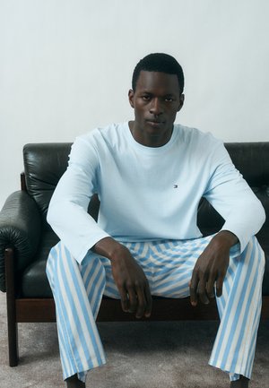 Mann trägt hellblaues Langarmhemd und blau-weiß gestreifte Hose, sitzt auf schwarzem Ledersofa vor einer schlichten Wand.