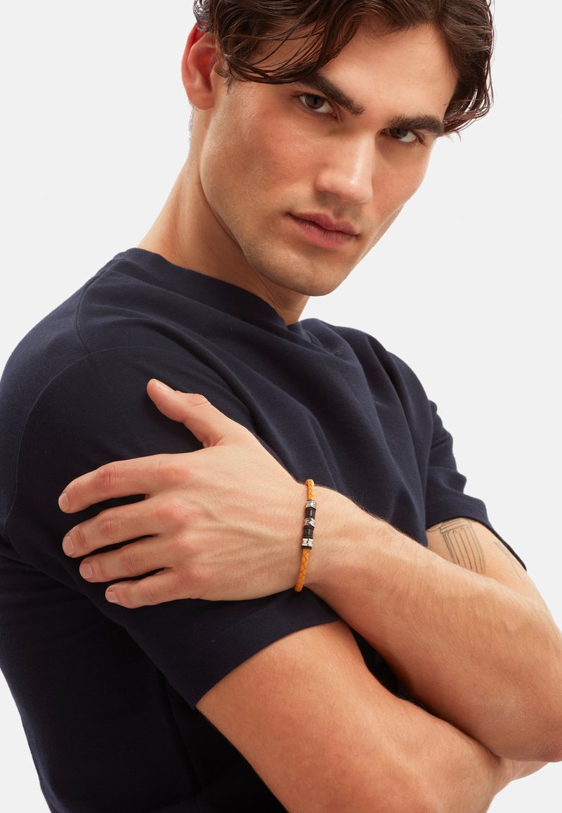 Pulsera de cuero trenzado naranja con acentos metálicos y tres cuentas negras distintas, llevada en una muñeca desnuda contra una camisa oscura.