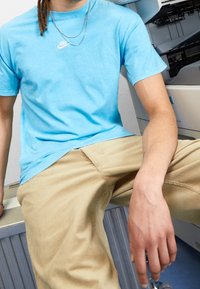 Personne portant un t-shirt Nike bleu et un pantalon beige assise près d'un équipement de bureau, montrant son torse et son bras avec des veines visibles.