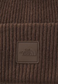 Brunt ribbestrikket stoff med et firkantet gummipatch preget med "THE NORTH FACE" logo i matchende brunt.