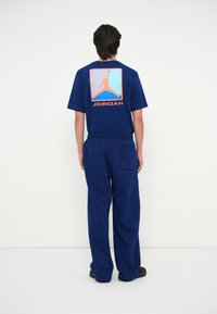 Marineblauw T-shirt met korte mouwen met een groot oranje Jumpman-logo op de achterkant en het woord "JORDAN" eronder. Gepaarde met bijpassende sweatpants.