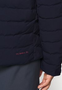 Marinblå puffjacka med quiltat mönster. Funktioner en röd "TX SHELL 2L"-etikett på fållen och kardborredetaljer på ärmen.