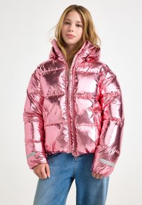 Teen girl wearing shiny metallic pink puffer jacket with hood and blue jeans, standing against plain white background.