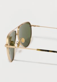 Óculos de sol aviador em metal dourado com lentes verdes, apresentando uma armação fina e o nome da marca gravado nas hastes, com um detalhe em tartaruga.