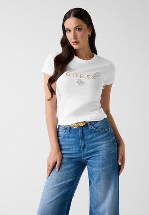 Kobieta o długich ciemnych włosach, ubrana w białą koszulkę z złotym napisem "GUESS" i motylkami, do tego niebieskie dżinsy i jasnobrązowy pasek.