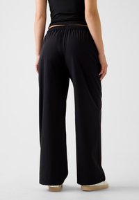 Pantalon noir ample avec taille élastique, texture lisse, jambes larges et détails de coutures subtils, associé à des chaussures de couleur claire.
